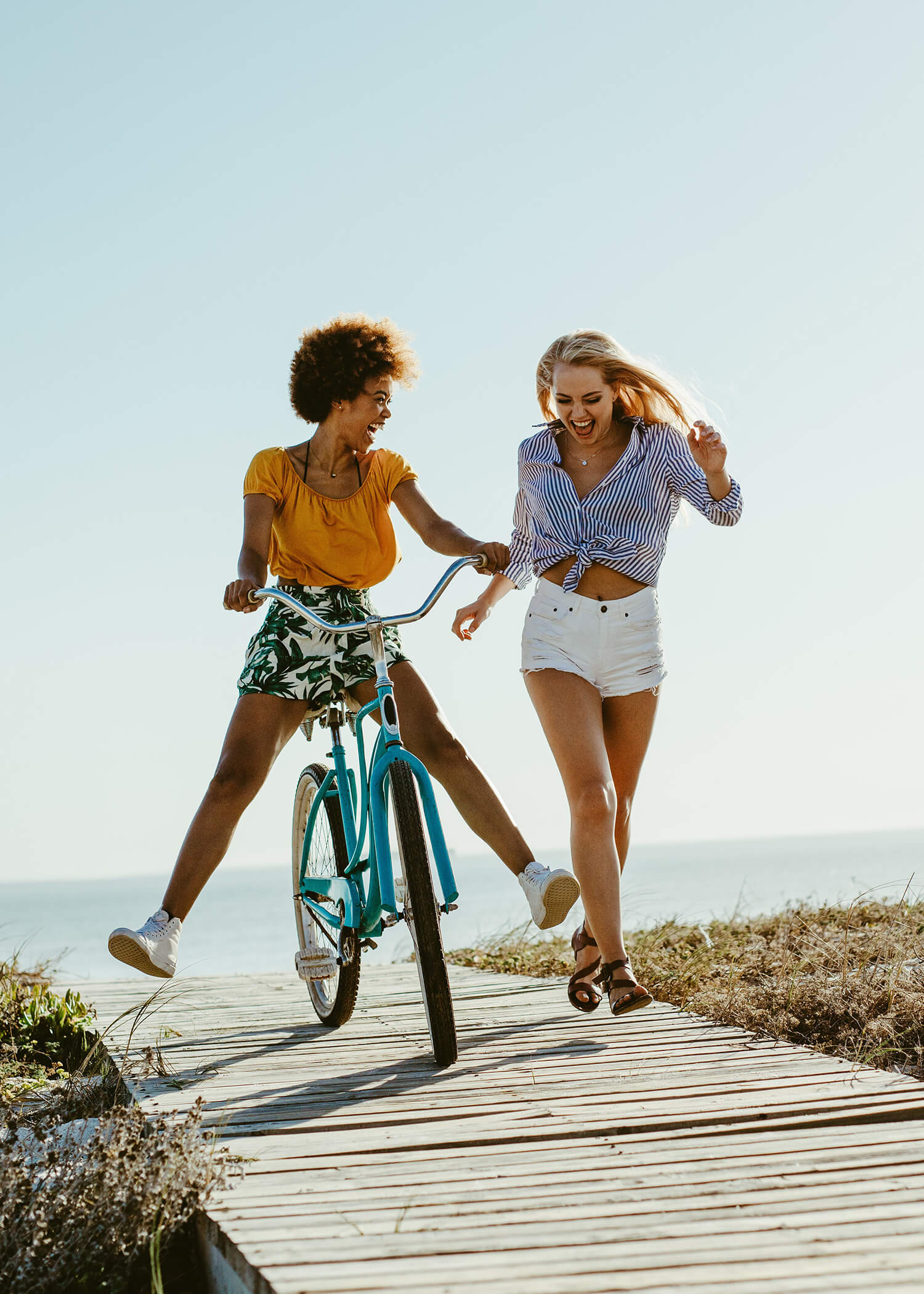 An image of two young women on a boardwalk smiling while one rides a bike. This image illustrates the full lives that people live after working with a DBT therapist in Torrance, CA through a DBT program. | 90274 | 90275