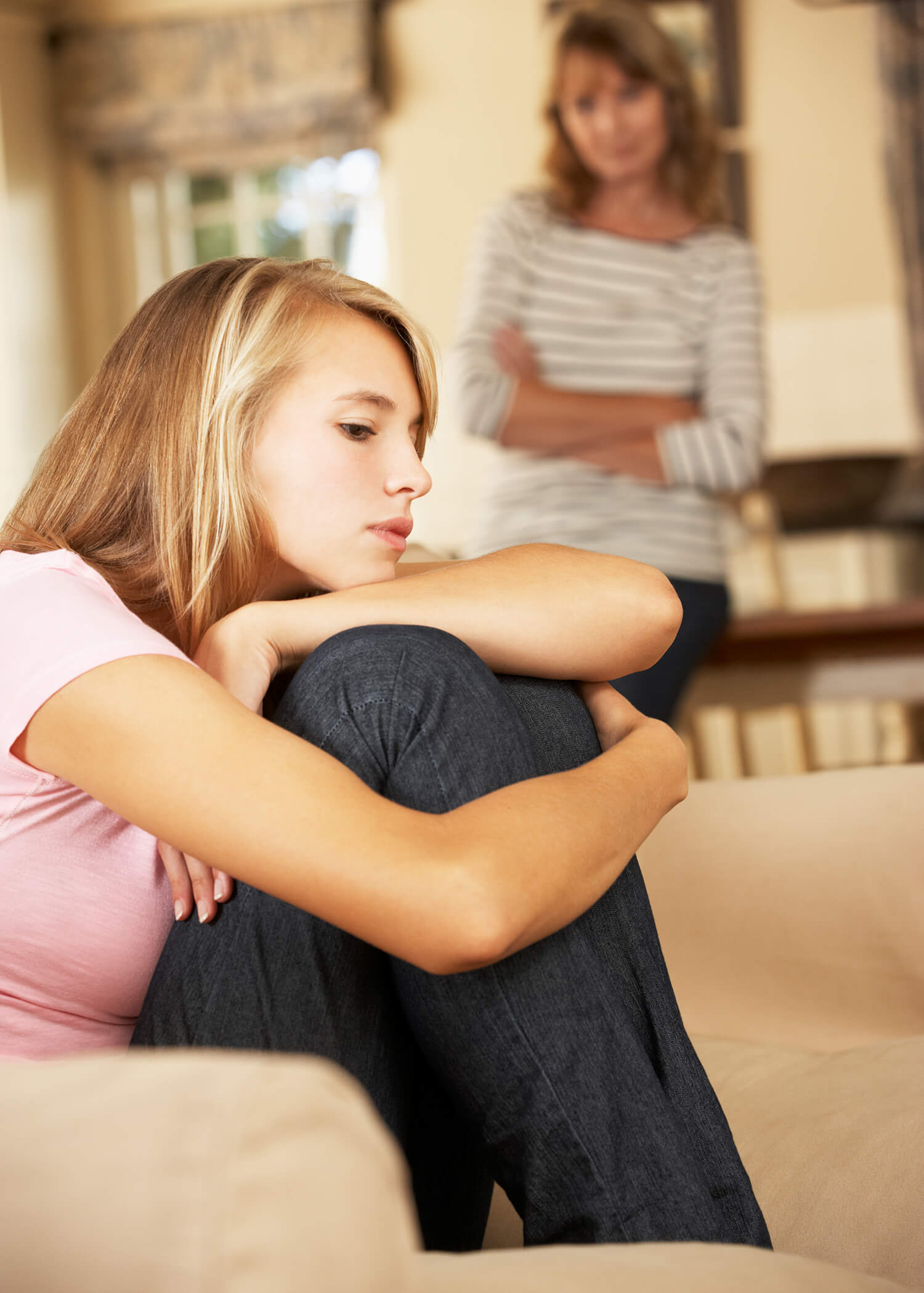 Image of a teen girl sitting with her knees to her chest and chin resting on them while her mother watches in the distance. Many adolescents encounter challenges for teens in Torrance, CA. Parents and teens can find support for family dynamics by working with a DBT therapist in Los Angeles, CA.  90254 | 90266