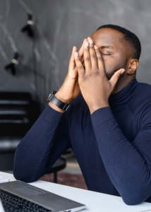 Image of a man sitting at a desk with his hands covering his face. This image depicts someone who is struggling with anger issues in Los Angeles, CA. People like this can learn about managing anger in Los Angeles, CA with DBT. | 90274 | 90275 | 90277