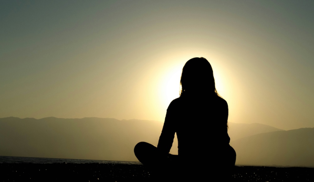 Image of a woman sitting crosslegged on the ground, staring at the sun as it rises. With mindfulness in Las Vegas, NV you can begin working toward coping with stress and anxiety in healthy ways.