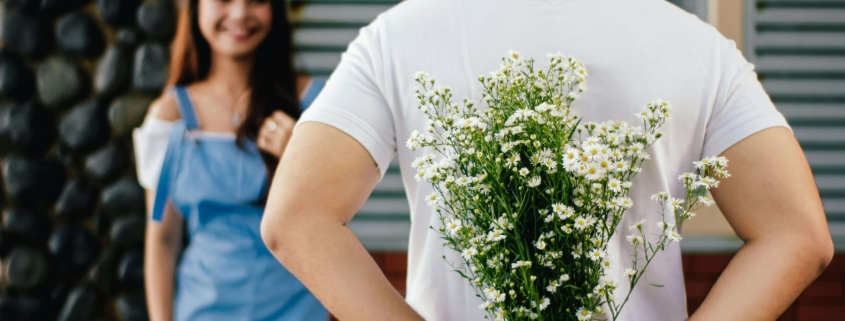 Shows a man in a white shirt holding white flowers behind his back in front of his wife. Represents how debt therapy in Las Vegas and a dbt therapist in Las Vegas can help you in your relationship.