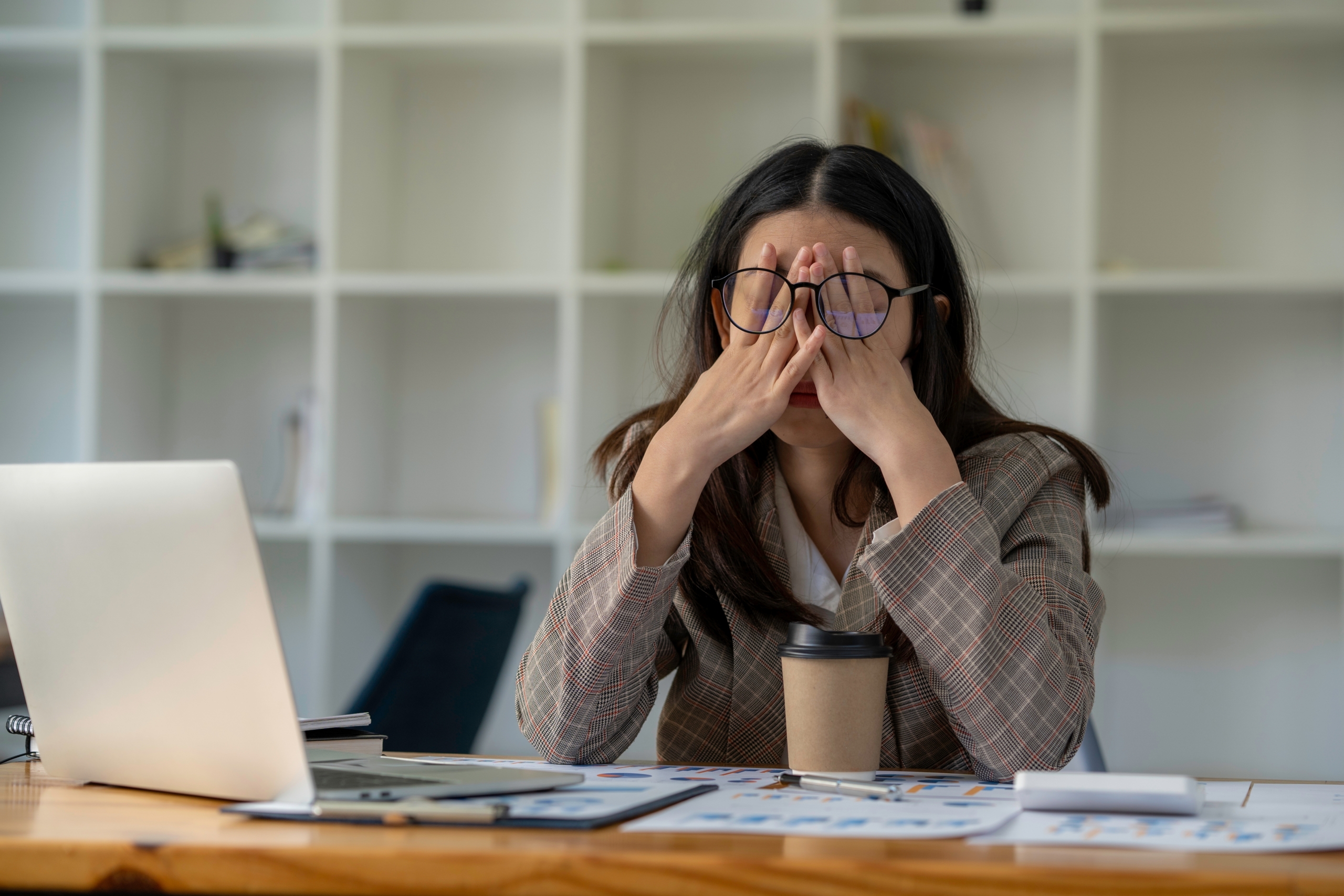 Overwhelmed woman holding her face while sitting at a desk with a laptop and coffee cup