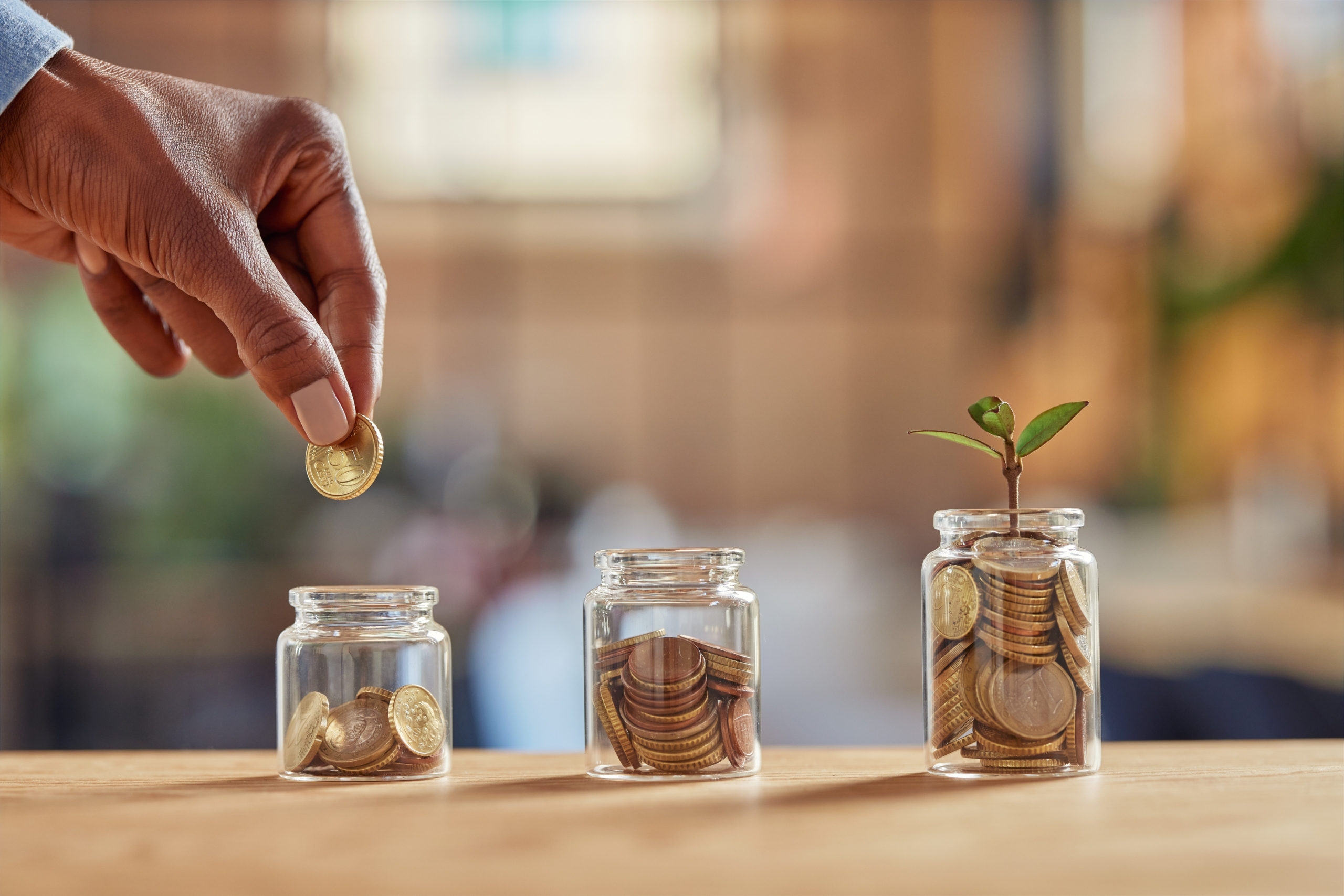 Hand placing a coin in one of three jars, each filling with coins, with a plant sprouting from the fullest jar — symbolizing DBT’s Accumulating Positives skill and emotional savings.
